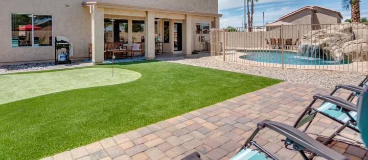 A backyard oasis in Sun Lakes AZ showcases a small putting green with premium artificial grass installation, a patio adorned with lounge chairs, and a pool boasting a waterfall. The house, with its large windows and doors, sits against palm trees and a clear sky—crafted by Maricopa Turf Pros.