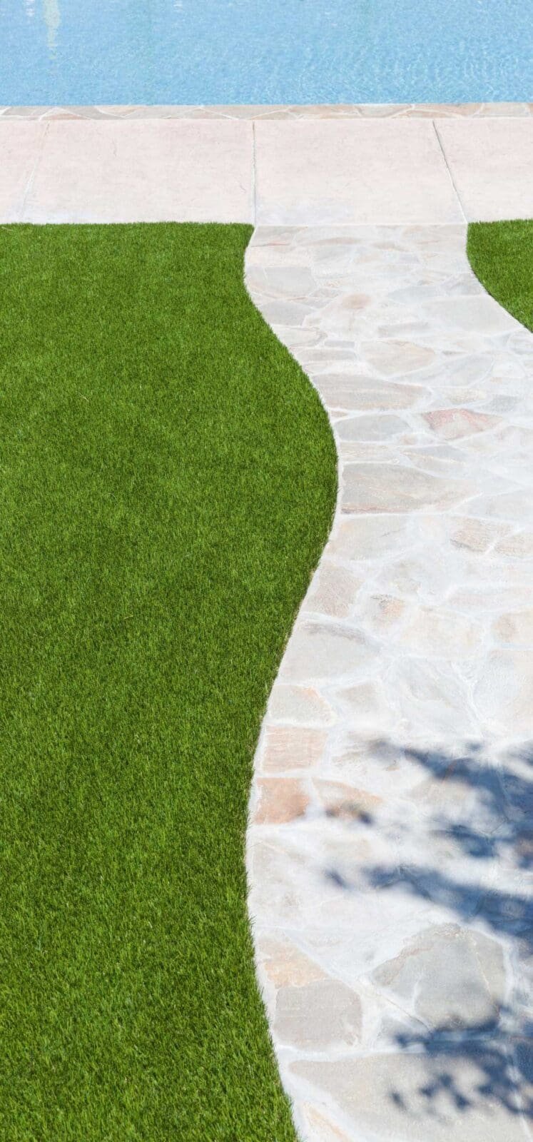 A curved stone pathway runs through a lush lawn of artificial grass, leading up to a sparkling blue swimming pool. This picturesque landscaping project in Maricopa AZ is bright and sunny, with the shadow of a tree partially visible on the grass.