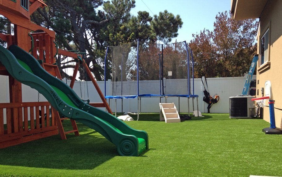 A backyard playground with a green slide attached to a wooden playset, a blue trampoline with a safety net, and a small swing set. The ground features playground turf installation by Maricopa Turf Pros. A white fence and trees are in the background.