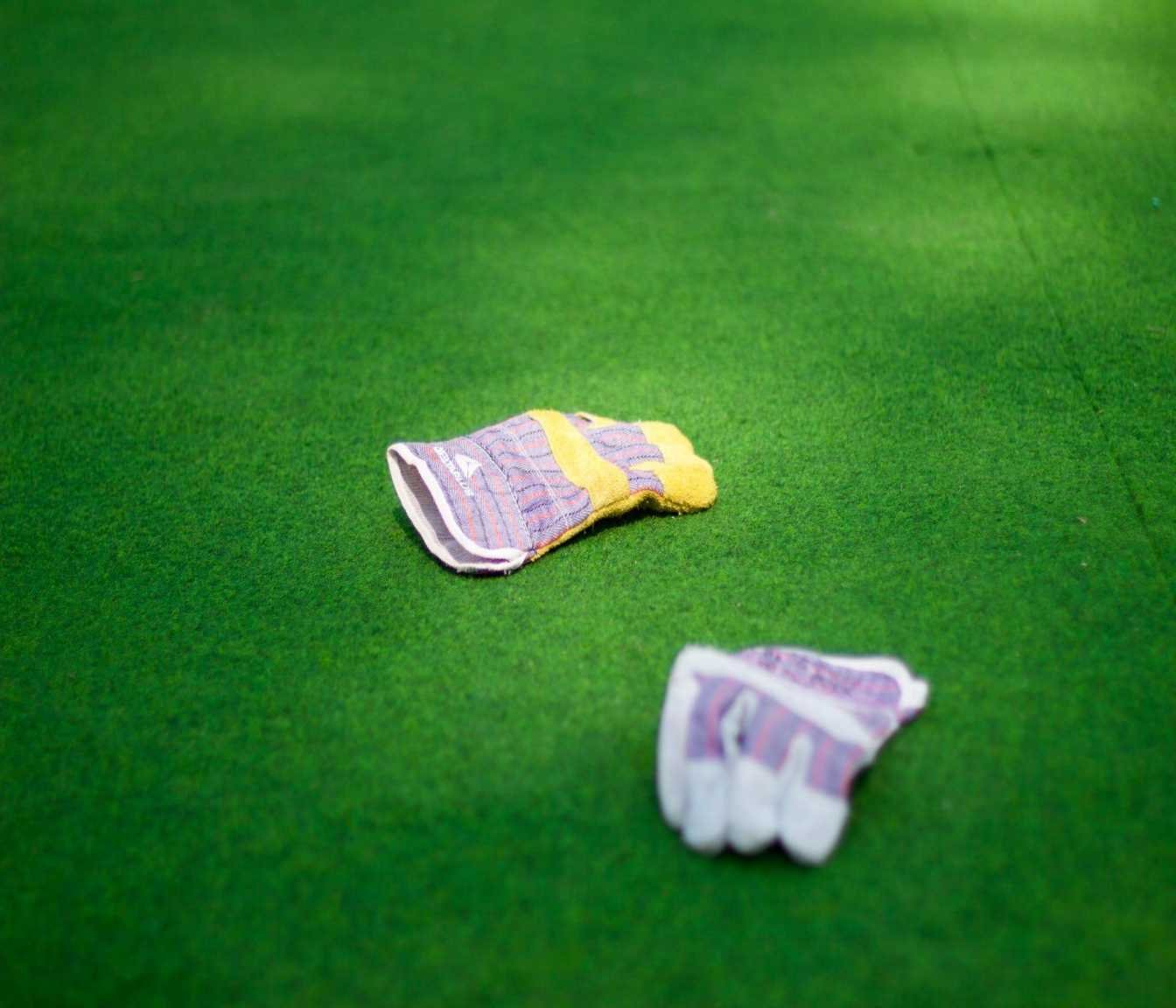 A pair of mismatched work gloves lies on a vibrant green lawn in Maricopa, AZ. One glove is predominantly yellow with pink and purple stripes, while the other glove is white with a similar pink and purple striped pattern. The gloves are positioned a distance apart from each other, perhaps after an athletic turf installation.