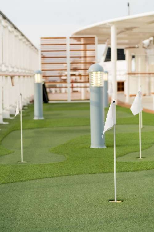 An outdoor mini-golf course on the deck of a cruise ship boasts expert turf installation with artificial green turf, small flags marking the holes, and modern lamp posts along the pathway. The ship's white railings and part of the structure are visible in the background, offering Free Quotes for installations.