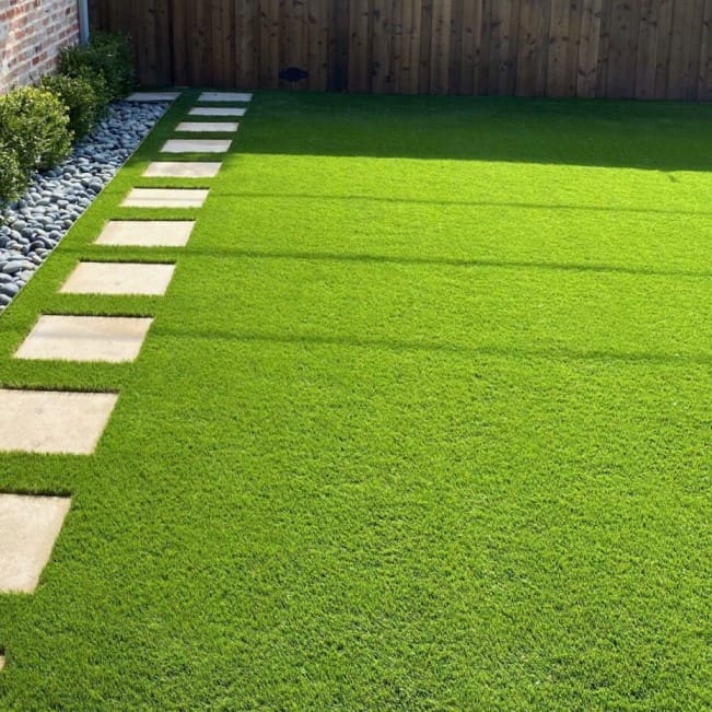 A well-maintained lawn with vibrant artificial grass, showcasing a row of rectangular stepping stones on the left. The lawn is bordered by decorative white stones and a row of green shrubs, reminiscent of Casa Grande elegance. A wooden fence stands proudly in the background.