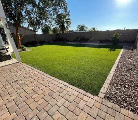 A backyard in Eloy, AZ, featuring a neatly manicured artificial grass lawn, surrounded by a brick-paved patio and gravel areas. A few trees and plants line the fenced perimeter under a clear blue sky.